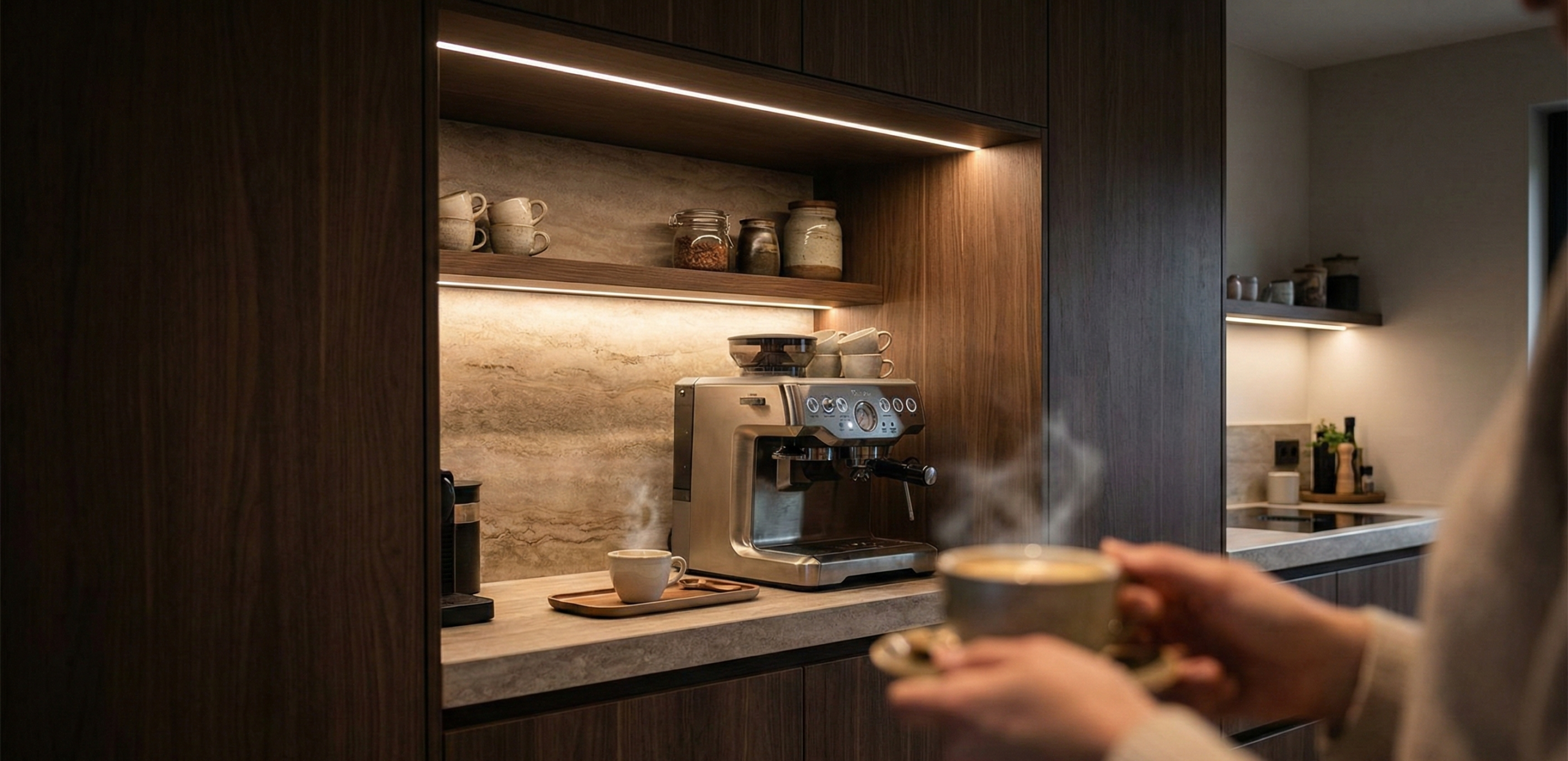 Persoon houdt een dampende kop koffie vast voor een luxe koffienis in de keuken, geïntegreerd in een donkere houten kastenwand met sfeervolle led-verlichting en een espressomachine.